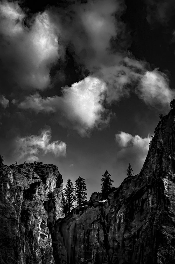 Majestic black and white landscape of rugged cliffs and towering pine trees under dramatic cloud-filled sky, artwork from The Doug Heslep Fine Art Collection.