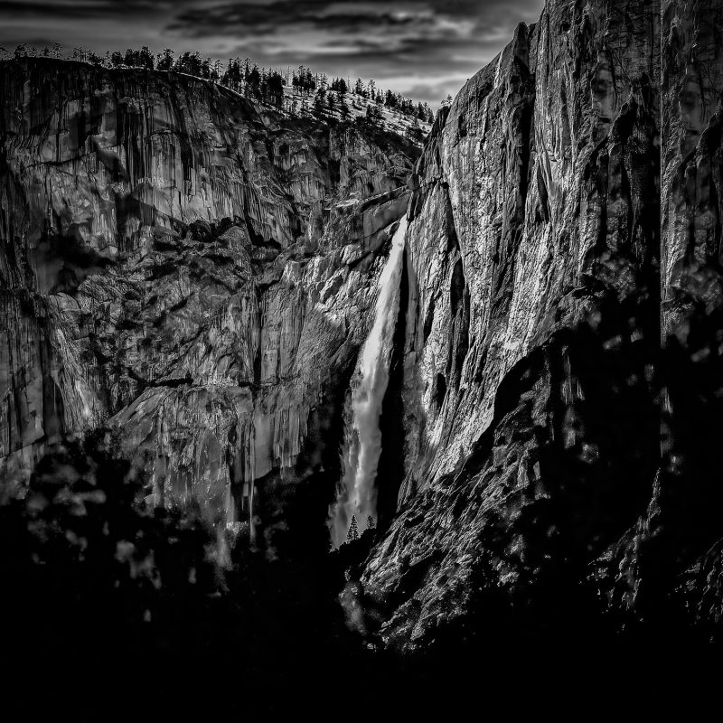 Moody black and white landscape photograph of Upper Yosemite Falls in Yosemite National Park, showcasing dramatic rocky cliffs and cascading waterfall.