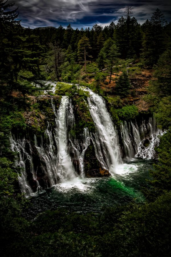 Raging Silver Cascade Waterfall amidst lush evergreen forest, under dark cloudy sky.