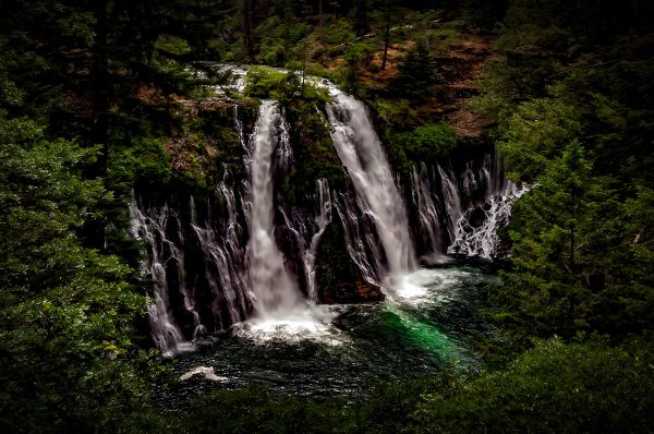 Majestic waterfall surrounded by lush green forest in the Doug Heslep Fine Art Collection.