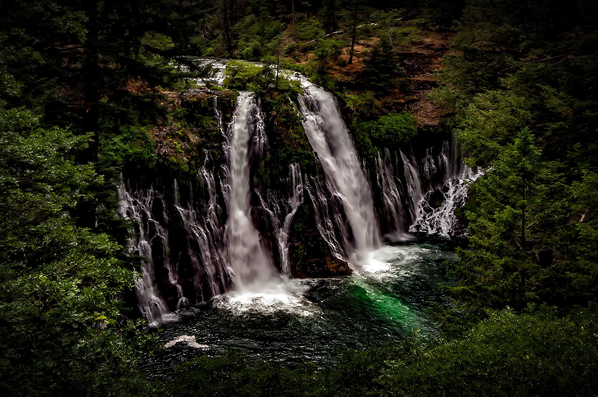Majestic waterfall surrounded by lush green forest in the Doug Heslep Fine Art Collection.