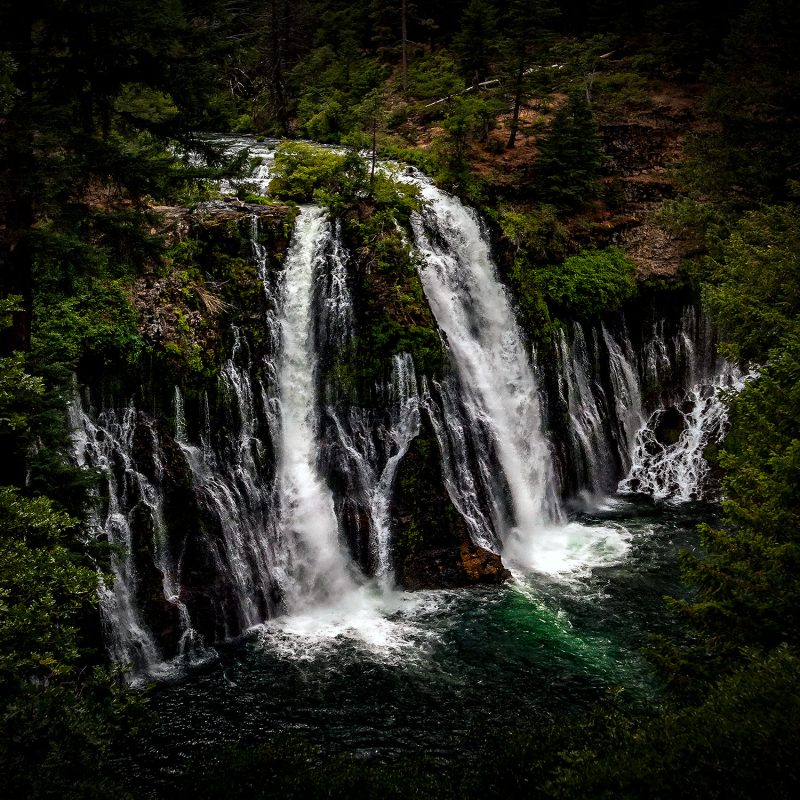 Serene waterfall surrounded by lush green trees in the Doug Heslep Fine Art Collection.