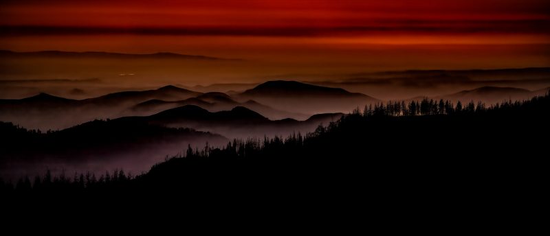 A stunning sunset over layered mountains with silhouetted trees, from The Doug Heslep Fine Art Collection.