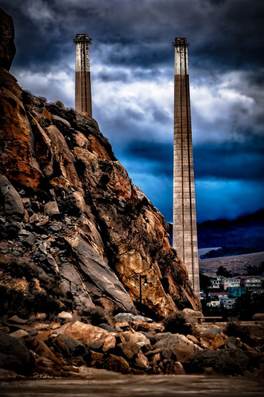 Chimney stacks in a rugged industrial landscape, evoke a nostalgic scene from The Doug Heslep Fine Art Collection.