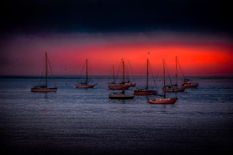 Boats anchored at sunset in the Doug Heslep Fine Art Collection, capturing vibrant sky colors and tranquil waters.