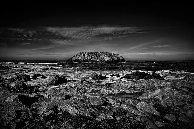 Rugged rocky coastline with large island in the distance, black and white seascape.
