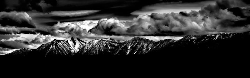 Snow-capped mountain range under dramatic cloudy sky in black and white.