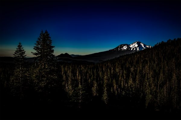Snow-capped mountain with dense evergreen forest under deep blue sky, part of The Doug Heslep Fine Art Collection.