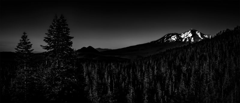 Dark mountain landscape in black and white, featuring tall trees and snow-capped peaks, from The Doug Heslep Fine Art Collection.