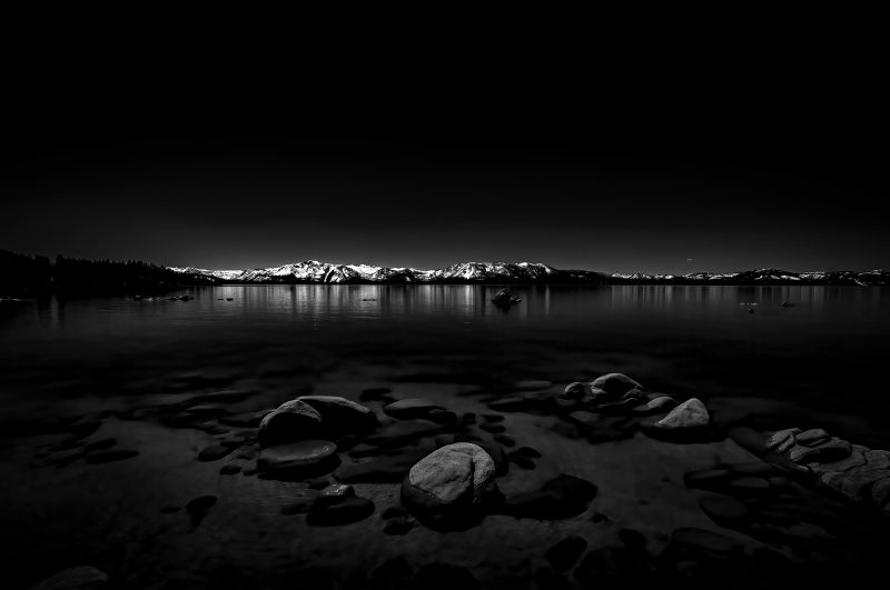 Black and white landscape photograph of snow-capped mountains reflecting on a calm lake with rocks in the foreground. Part of The Doug Heslep Fine Art Collection.