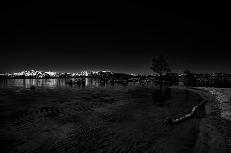 Dark landscape photograph of snow-capped mountains and trees reflected in calm lake water.