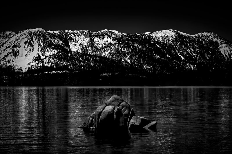 Wild mountain landscape with snow-capped peaks, reflective lake, and rocks in black and white.