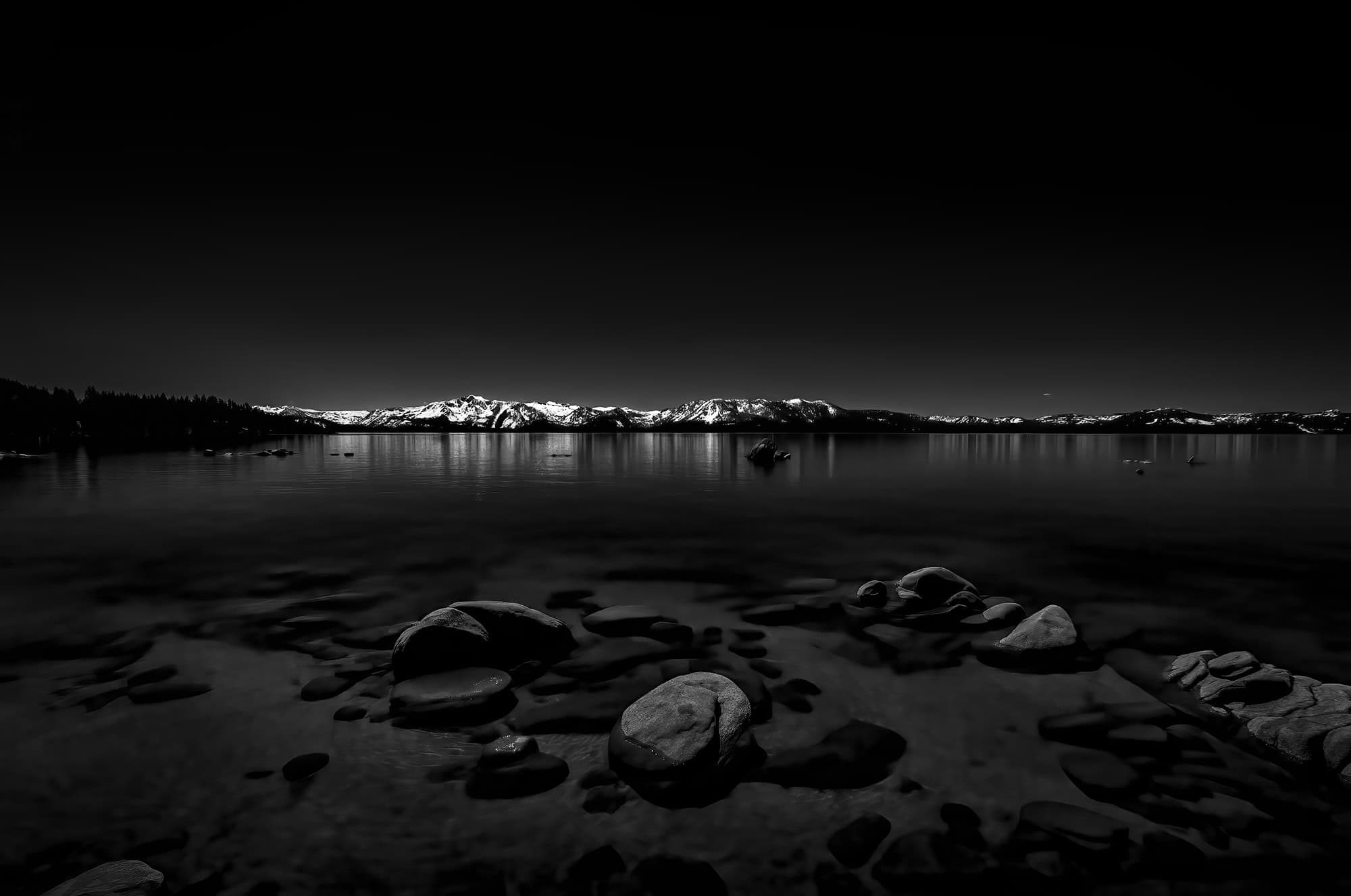 Black and White photo looking across Lake Tahoe with rocks in the foreground and snow covered mountains in the background