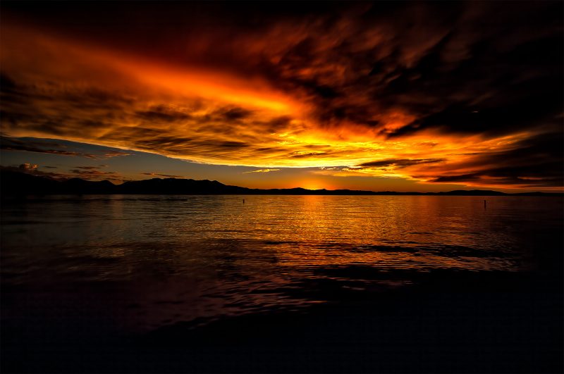 Vibrant sunset over calm lake with dramatic cloud formations and mountain silhouette.