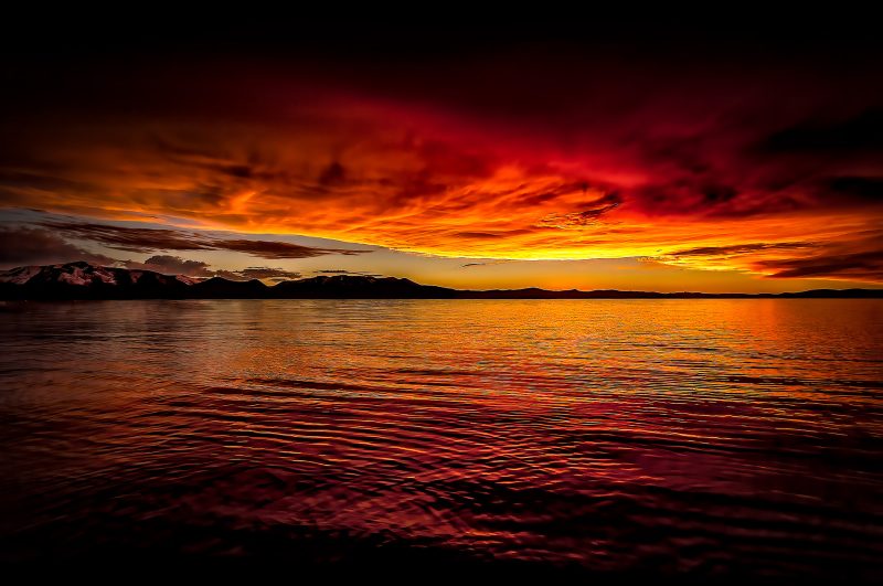 Vivid sunset over a tranquil lake with mountains in the background, showcasing dramatic clouds and fiery colors.