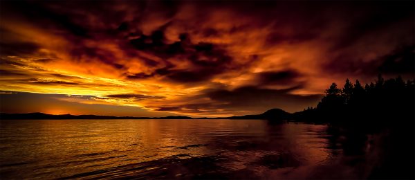 Vivid sunset over a tranquil lake with dramatic orange and purple clouds and silhouetted trees.