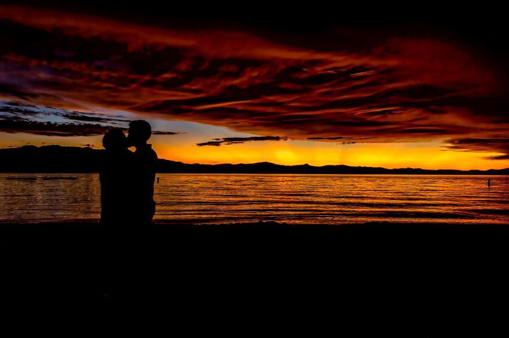 Silhouetted couple embracing at sunset by the water, vibrant orange and red sky, a serene romantic scene.