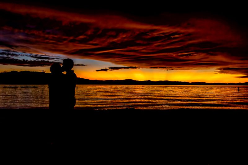 Silhouetted couple embracing at sunset by the water, vibrant orange and red sky, a serene romantic scene.