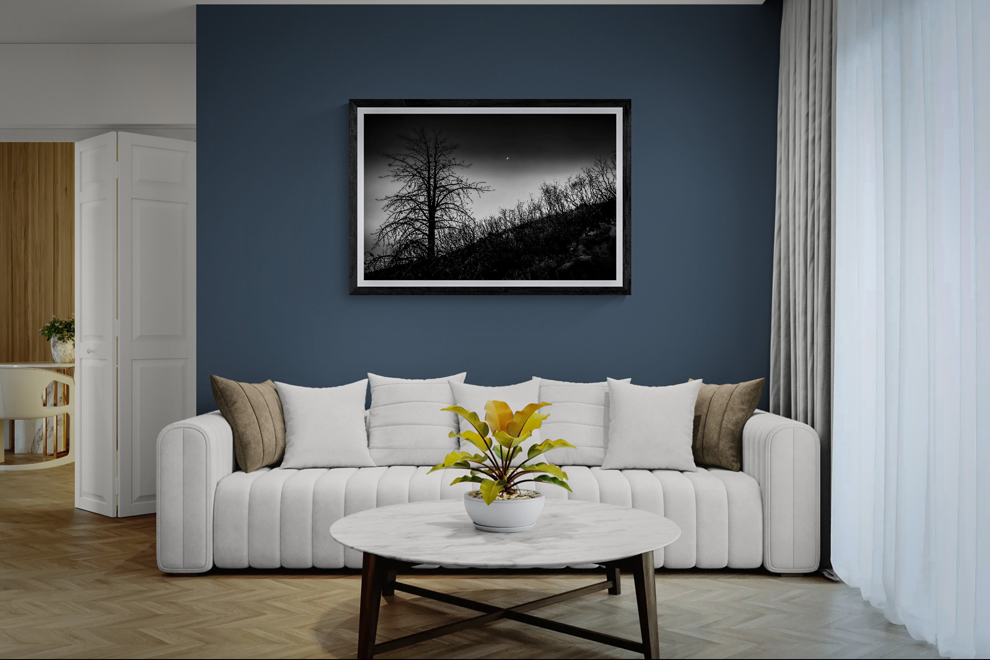 A black and white landscape photograph of a tree on a hill, framed and hung on a blue wall in a mode.