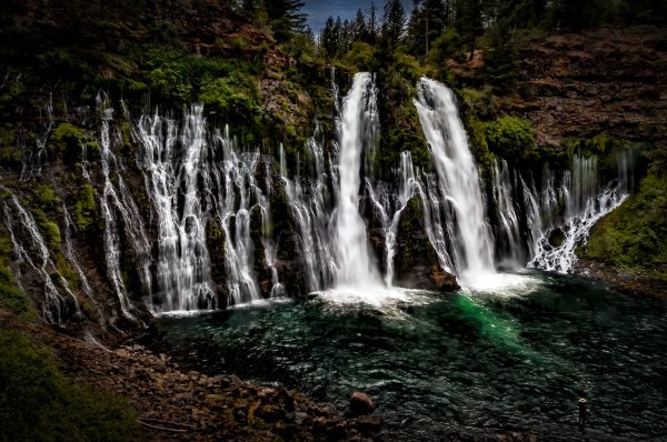 A full view of Burney Falls from halfway down the descending trail showing a lone fisherman casting a line.